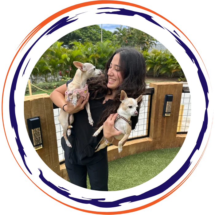 A smiling woman in black clothing holding two small white dogs outdoors near a fenced area.